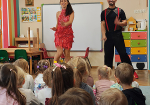 Przedszkolaki siedzą na dywanie, przed nimi kobieta w czerwonej sukience i mężczyzna w czarnym ubraniu.