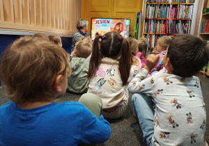 Przedszkolaki siedzą na dywanie. Przed nimi siedzi Pani bibliotekarka - pokazuje im na ekranie program edukacyjny o jesieni.