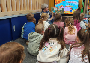 Przedszkolaki siedzą na dywanie. Przed nimi siedzi Pani bibliotekarka - czyta im książkę o jesieni.