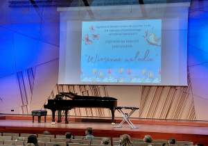 Scena sali koncertowej Szkoły Muzycznej. Na scenie stoi fortepian i krzesełko. Na ścianie wisi tablica interaktwna z wyświetlonym tytułem koncertu.