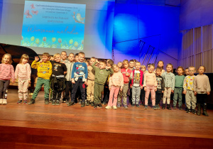 Dzieci stoją na scenie sali koncertowej Szkoły Muzycznej. Za nimi stoi fortepian. Na ścianie wisi tablica interaktwna z wyświetlonym tytułem koncertu.