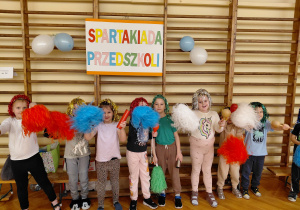 Przedszkolaki przebrane w świecące peruki, z pomponami i instrumentami stoją na parkiecie sali gimnastycznej. Za nimi na drabinkach wisi napis: Spartakiada Przedszkoli.