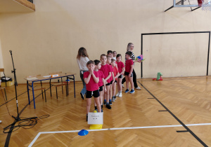Dzieci w czerwonych koszulkach stoją w rzędzie na sali gimnastycznej. Za nimi idzie dziwczynka i kobieta. Obok stoi ławka, krzesła i stojak na mikrofon.