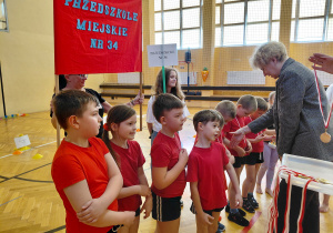 Przedszkolaki w czerwonych koszulkach stoją w szeregu. Część dzieci ma medale. Przed nimi stoi kobieta z wyciagniętą dłonią. Za dziećmi stoi kobieta z szyldem Przedszkola Miejskiego nr 34. Obok kobiety stoi dziwczynka z szyldem Przedszkola Miejskieg nr 34.