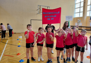 Przedszkolaki w czerwonych koszulkach i medalach stoją w szeregu. Jeden z chłopców trzyma puchar. Za dziećmi stoi kobieta z szyldem Przedszkola Miejskiego nr 34. Obok kobiety stoi dziwczynka z szyldem Przedszkola Miejskieg nr 34.