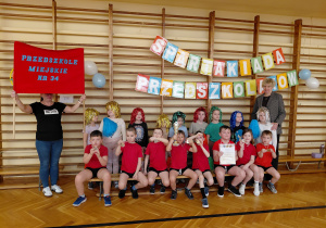 Przedszkolaki przebrane w świecące peruki, z pomponami stoją przed drabinkami. Przed nimi na ławce siedzą dzieci w czerwonych koszulkach z medalami. Jeden z chłopców trzyma puchar, drugi z chłopców trzyma dyplom. Obok dzieci stoi kobieta w marynarce. Z drugiej strony stoi kobieta z szyldem Przedszkola Miejskiego nr 34. Za nimi, na drabinkach wisi napis Spartakiada Przedszkolaków.