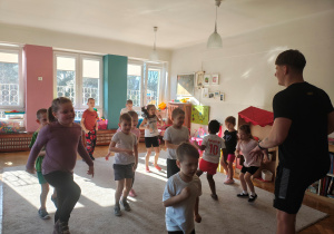 Przedszkolaki biegną w miejscu na dywanie. Mężczyzna biegnie w miejscu przodem do dzieci.