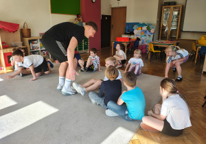 Część dzieci siedzi w rzędzie na dywanie. Na środku dywanu stoi pochylony mężczyzna. Pozostałe dzieci siedzą na krańcu dywanu.