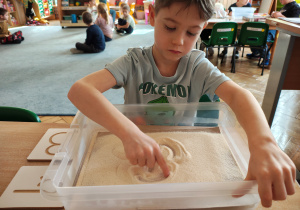Chłopiec siedzi przy stole i pisze litery palcem po kaszy mannie w pudełku. Obok pudełka leżą litery. W tle dzieci siedzące na dywani i przy stole.