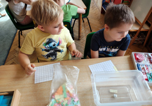 Dwóch chłopców siedzi przy stole. Przed nimi leżą kartki w dużą kratę, kolorowe kartoniki oraz pudełko. Jeden z chłopcow trzyma kostkę do gry.