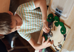 Chłopiec siedzi przy stole i układa drewnianą układankę drzewa.