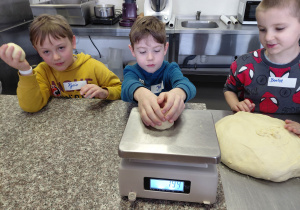 Chłopiec waży surowe ciasto. Obok niego stoją chłopcy z surowym ciastem.