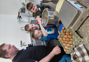 Chłopiec wbija jajko do misy na ciasto. Obok niego stoi dwóch chłopców i mężczyzna.