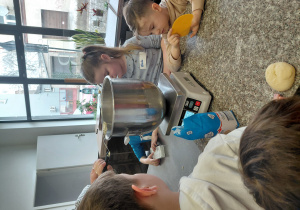 Dzieci stoją przy stole z wagą, na której stoi misa.