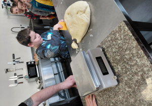 Chłopiec odcina kawałek surowego ciasta. W tle rząd dzieci czekających na swoją kolej.