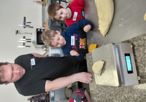 Przy stole stoi mężczyzna i dwóch chłopców. Na stole stoi waga. Na wadze leży kawalek surowego ciasta.