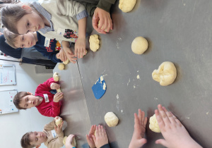 Przedszkolaki stoją przy stole i formują bułki z surowego ciasta.