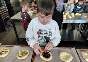Chłopiec dekoruje uformowaną bułkę truskawkami. W tle stoją dzieci przy stole.