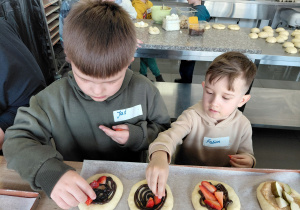 Dwóch chłopców dekoruje uformowane bułki truskawkami. W tle stoją dzieci przy stole.
