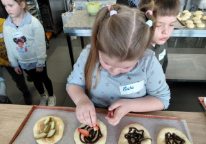 Dziewczynka dekoruje uformowaną bułkę truskawkami. Za nią stoją dzieci.