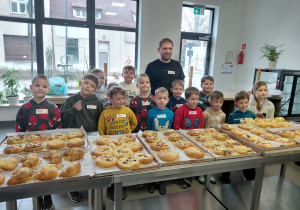 Przedszkolaki wraz z mężczyzną stoją za stołem, na którym leżą upieczone bułki drożdżowe.