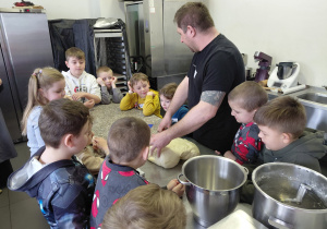 Przedszkolaki wraz z mężczyzną stoją przy stole. Na stole leży surowe ciasto.