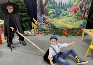 Chłopiec w przebraniu czarownicy trzyma łopatę, na której siedzi drugi chłopiec. W tle obrazek lasu i chatki.