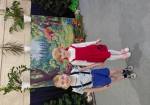 Chłopiec i dziewczynka stoją na dywanie. W tle obrazek lasu i chatki.