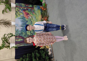 Chłopiec i dziewczynka stoją na dywanie. W tle obrazek lasu i chatki.