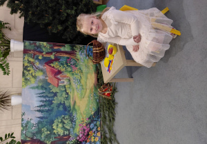 Dziewczynka siedzi na krześle przy stoliku. W tle obrazek lasu i chatki.