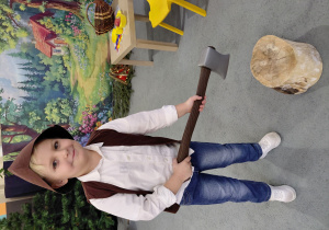 Chłopiec w przebraniu drwala, trzyma zabawkową siekierę. W tle obrazek lasu i chatki.