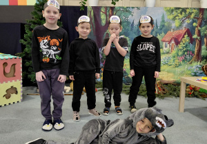 Czterech chłopców w czarnych strojach, z obrazkami wilka na głowach stoi na dywanie. Jeden chłopiec w przebraniu wilka leży na dywanie. W tle obrazek lasu i chatki.