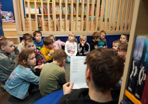 Przedszkolaki siedzą w kole na dywanie. Starszy chłopiec siedzi tyłem i czyta zagadki. W tle regały z książkami.