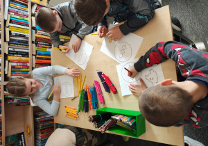 Trzech chłopców i jedna dziewczynka siedzą przy stole i kolorują kurczaczka i pisankę.