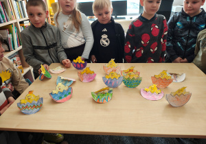 Przedszkolaki stoją za stołem, na którym stoją papierowe kurczaczki w pisankach.