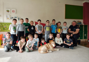 Przedszkolaki siedzą i stoją na dywanie, obok na jednym kolanie klęczy mężczyzna. Przed dziećmi leży pies.