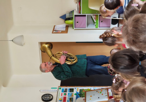 Mężczyzna gra na sakshornie barytonowym. Przed nim siedzą dzieci.