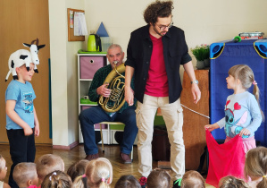 Mężczyzna gra na sakshornie barytonowym siedząc na krześlę. Przed nim dwoje dzieci - człopiec w czapce krowy oraz dziewczynka z czerwoną chustą. Pomiędzy dziećmi stoi mężczyzna w okularach.