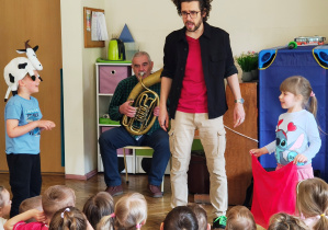 Mężczyzna gra na sakshornie barytonowym siedząc na krześlę. Przed nim dwoje dzieci - człopiec w czapce krowy oraz dziewczynka z czerwoną chustą. Pomiędzy dziećmi stoi mężczyzna w okularach.