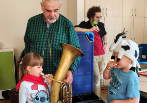 Dziewczynka w czerwonej chuście gra na sakshornie barytonowym, chłopiec w czapce krowy przykłada ustnik do ust. Mężczyzna w koszuli w kratę przytrzymuje instrument dziewczynce. Za nimi przy leżakach stoi mężczyzna w okularach.