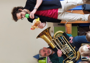 Mężczyzna w koszuli w kratę trzyma tubę siedząc na krześle. Obok stoi mężczyzna w okularach trzymając pluszową kurę.