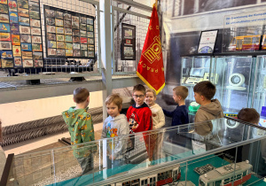 Dzieci stoją przy fladze MPK. Przed nimi modele starych tramwajów.