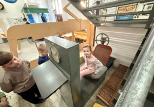 Dziewczynka siedzi na starym krześle przy maszynie do sprawdzania siły nóg. Obok maszyny stoi dwóch chłopców.