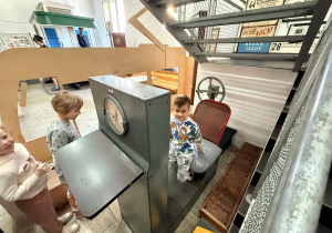 Chłopiec siedzi na starym krześle przy maszynie do sprawdzania siły nóg. Obok maszyny stoją dwie dziewczynki.