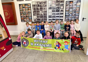 Przedszkolaki siedzą na starej ławce tramwajowej oraz przed nią. Tzrymają baner Akademii Kulturalnego Pasażer. Za nimi zdjęcia związane z łódzką komunikację miejską.