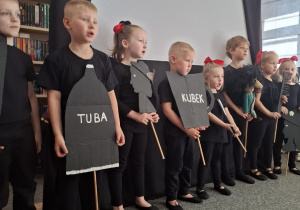 Przedszkolaki ubrane na czarno stoją przed białym ekranem.