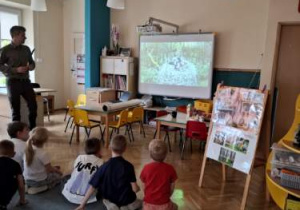 Przedszkolaki siedzą na dywanie przodem do tablicy multimedialnej, leśniczy stoi przy biurku.