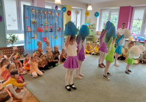 Dziewczynki stoją na dywanie trzymając pompony w górze. Pozostałe dzieci siedzą na podłodze.