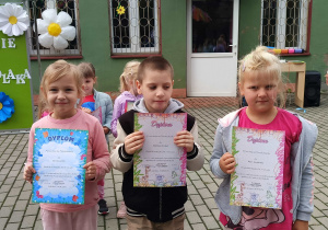 Dziewczynki i chłopiec stoją z dyplomami na tarasie przedszkolnym.