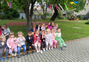 Przedszkolaki siedzą na ławkach i machają rączkami.